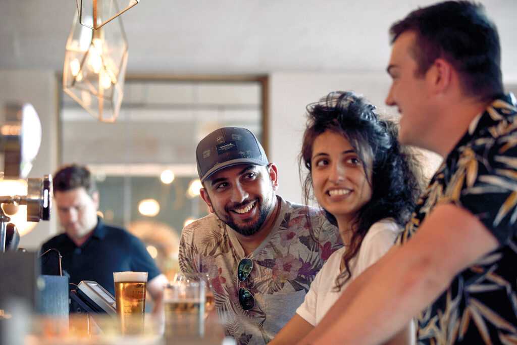 Three friends are chatting while drinking at the bar. In the background, a large mirror reflects the room.