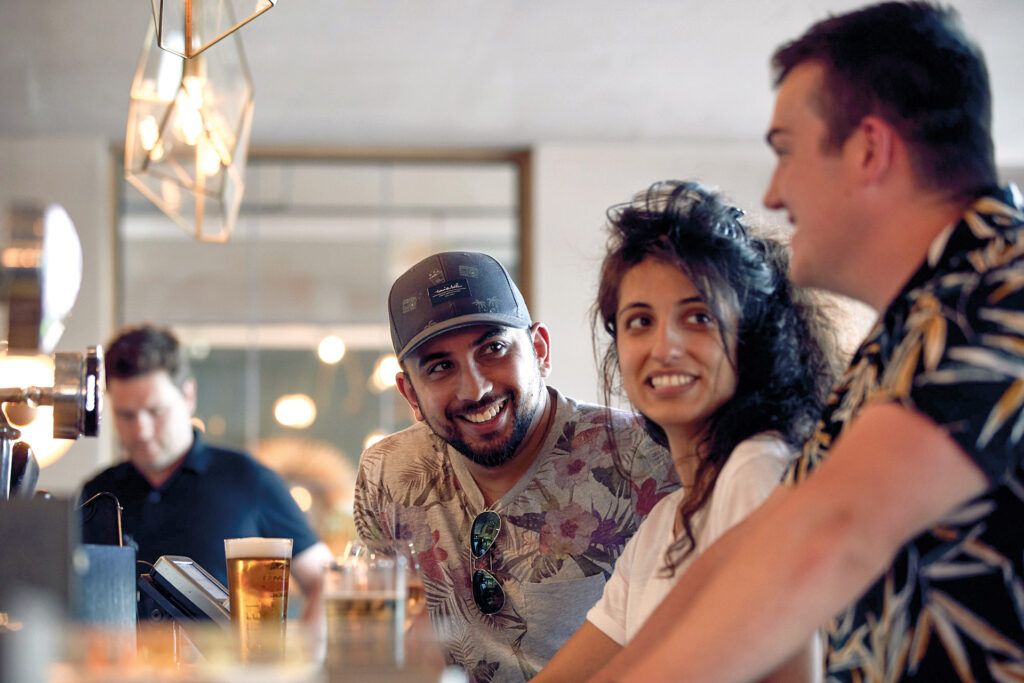 Three friends are chatting while drinking at the bar. In the background, a large mirror reflects the room.