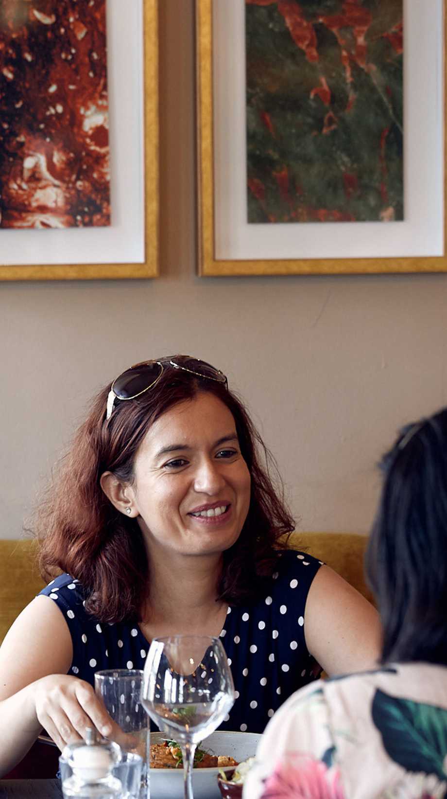 A woman in a polka dot top with sunglasses on her head talks to her friend, sat opposite her at a table.