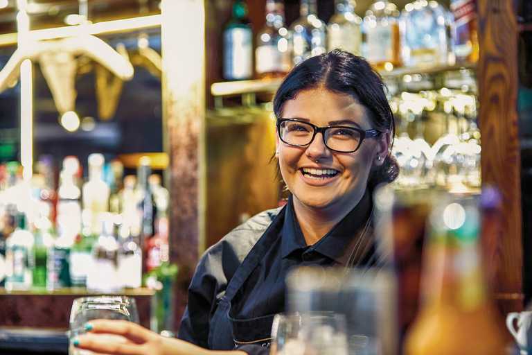 A team member smiles at the camera, holding a glass in their hand. Behind her, the bar twinkles with glasses and bottles.