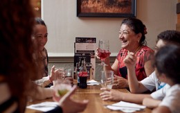 A family sit around a table together, laughing and sharing drinks. One woman is holding a fruity cider with ice.