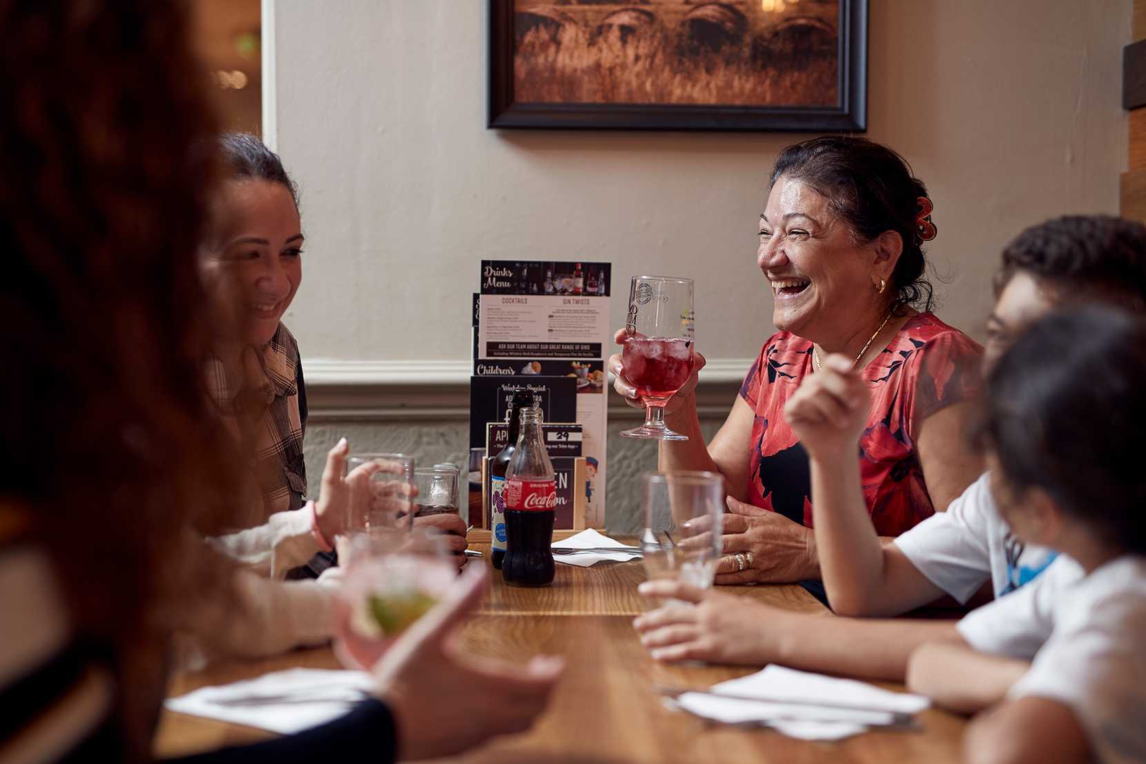 A family sit around a table together, laughing and sharing drinks. One woman is holding a fruity cider with ice.