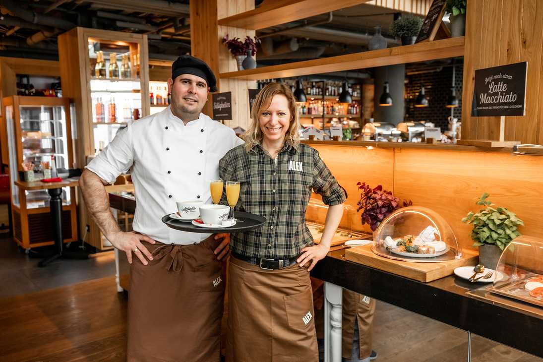 A chef and front-of-house team member are waiting at the door for guests, holding a tray with drinks.