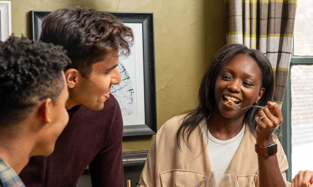 Three friends are having a conversation. Two men look over at a smiling woman as she raises a fork to her mouth.