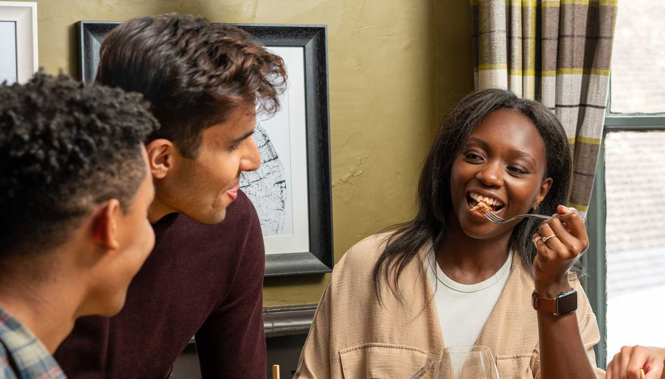 Three friends are having a conversation. Two men look over at a smiling woman as she raises a fork to her mouth.