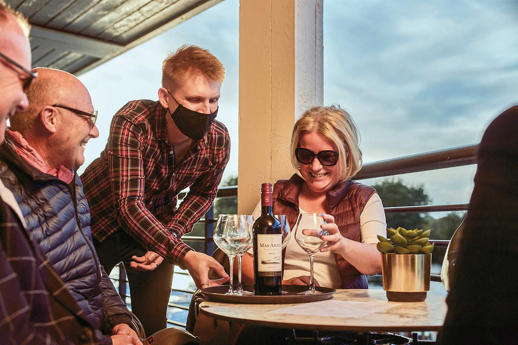 A team member in a checkered shirt and face mask delivers a bottle of red wine and four glasses to a table of smiling guests.