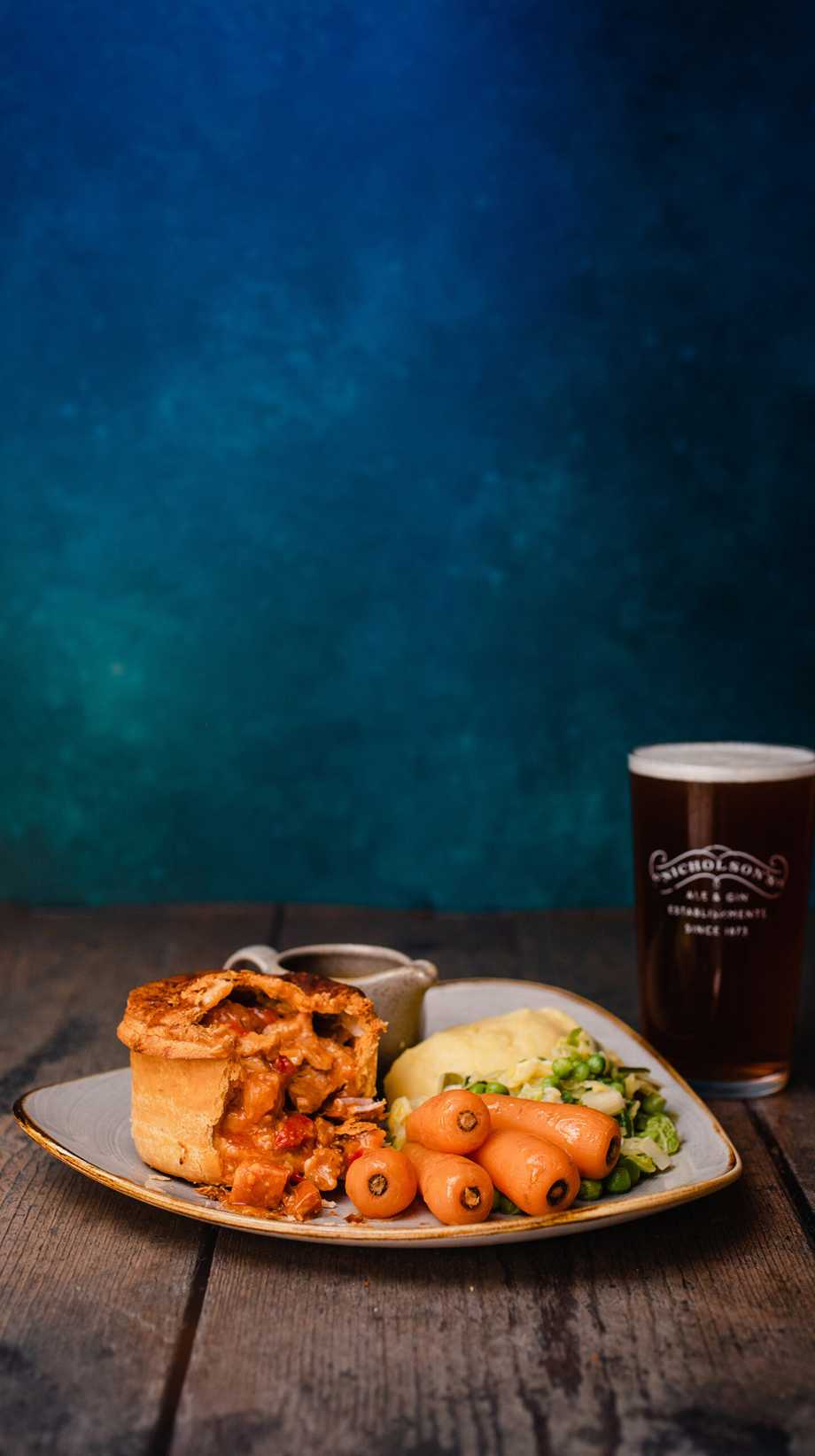 A plate sits against a blue background. On it, there's a pie, mash, vegetables, and gravy. Behind it, a pint of Nicholson's ale.