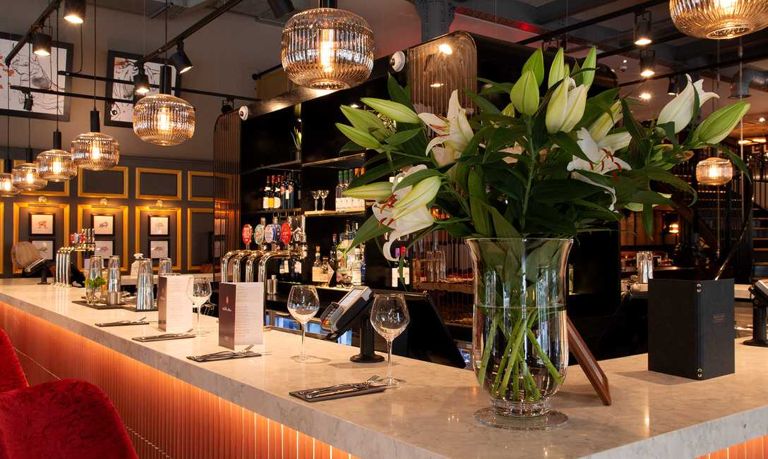 In Miller & Carter Cardiff, wine glasses, menus, and cutlery line the marble bar. On the corner of the bar, fresh lilies bloom in a glass vase and soft lighting hangs from the ceiling.