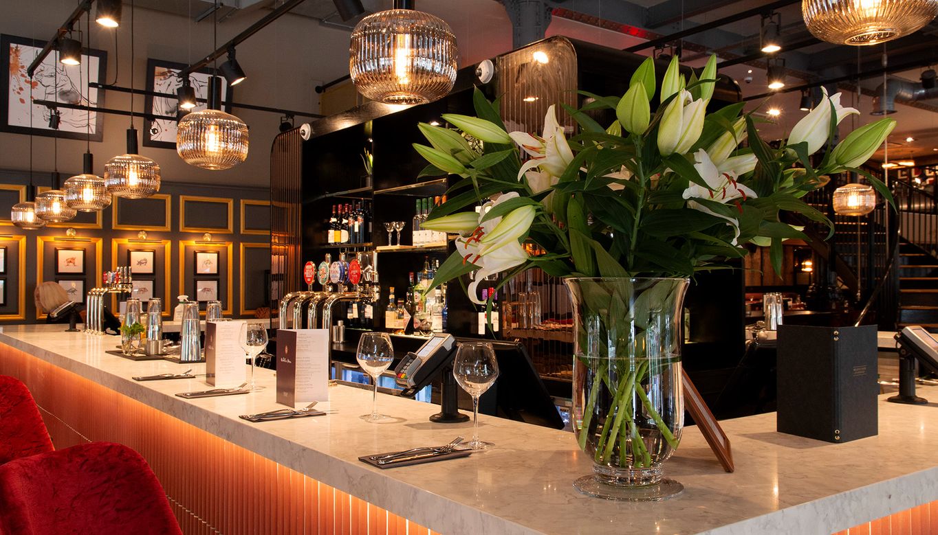 An interior shot of Miller & Carter Cardiff, showing the bar seating area decorated with lilies, soft lighting, and menus.