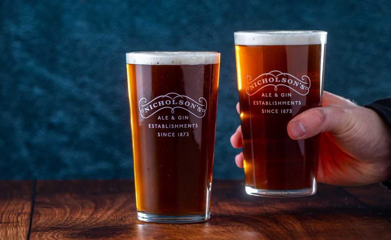 Two pints of Nicholson's Ale against a blue background. One pint is being picked up.