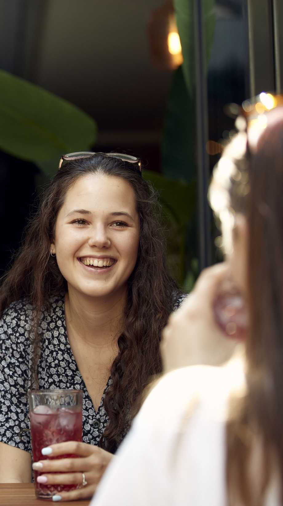 A girl with glasses resting on her head smiles at her friend, sitting in a tastefully decorated bar with plants and exposed lightbulbs.