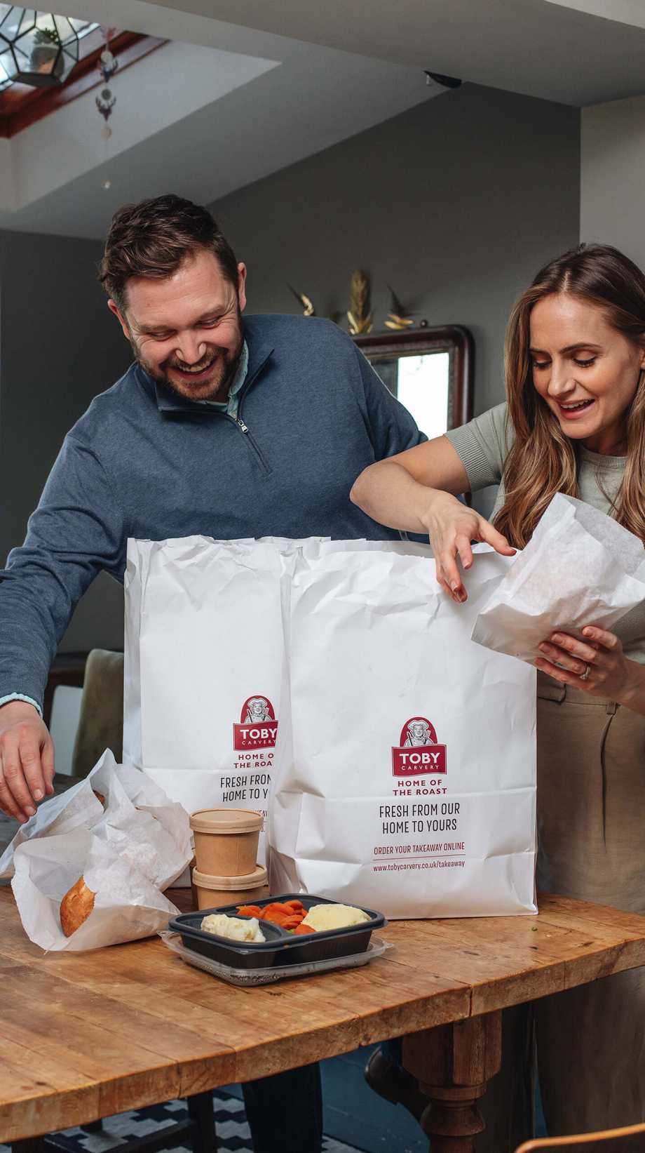 A couple place a Toby Carvery takeaway on their table at home. Meat, veggies, and mash are neatly packed in front of tubs of gravy.