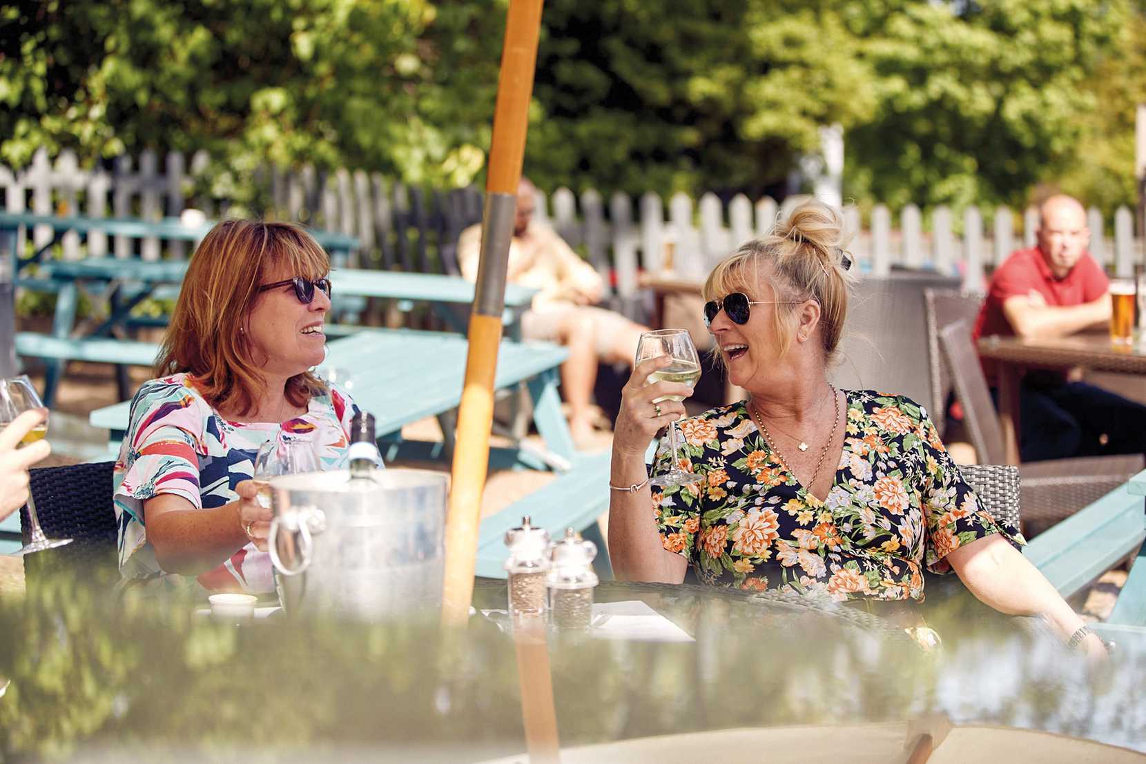Two woman wearing sunglasses are drinking in the sun. In the background, a man drinks a beer.