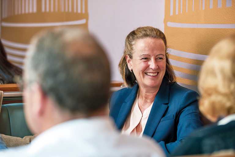 A woman wearing a dark blue suit sits in a booth, smiling at her colleagues.