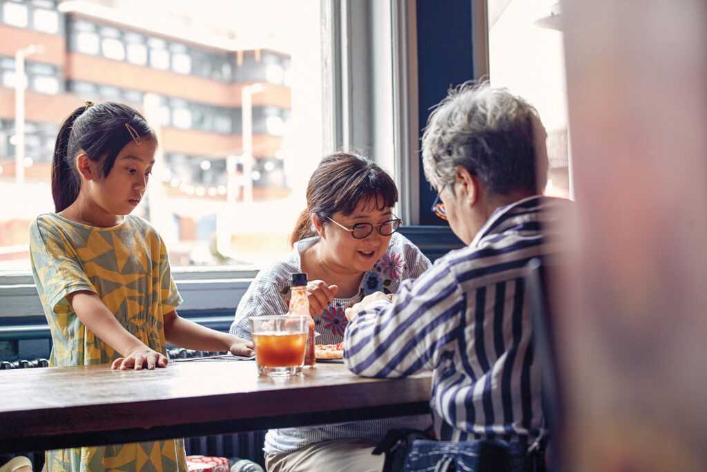 A family share a pizza and drinks at a window seat. The young girl stares at the pizza in awe.