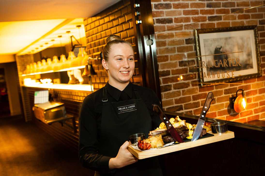 A team member carries a wooden block with steak, sides, and sauce through Miller & Carter.