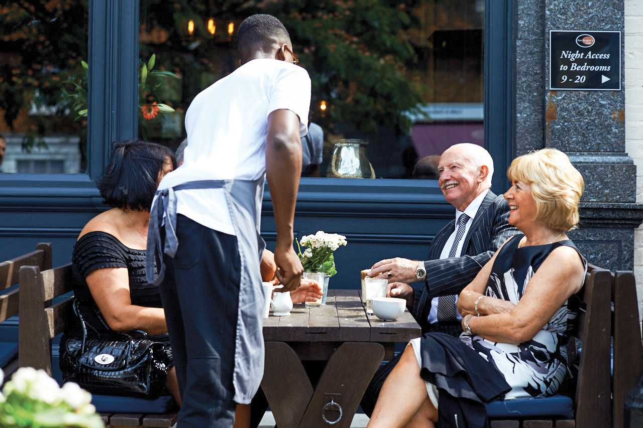 A team member wearing an apron serves drinks to a table of older guests, dressed smartly.