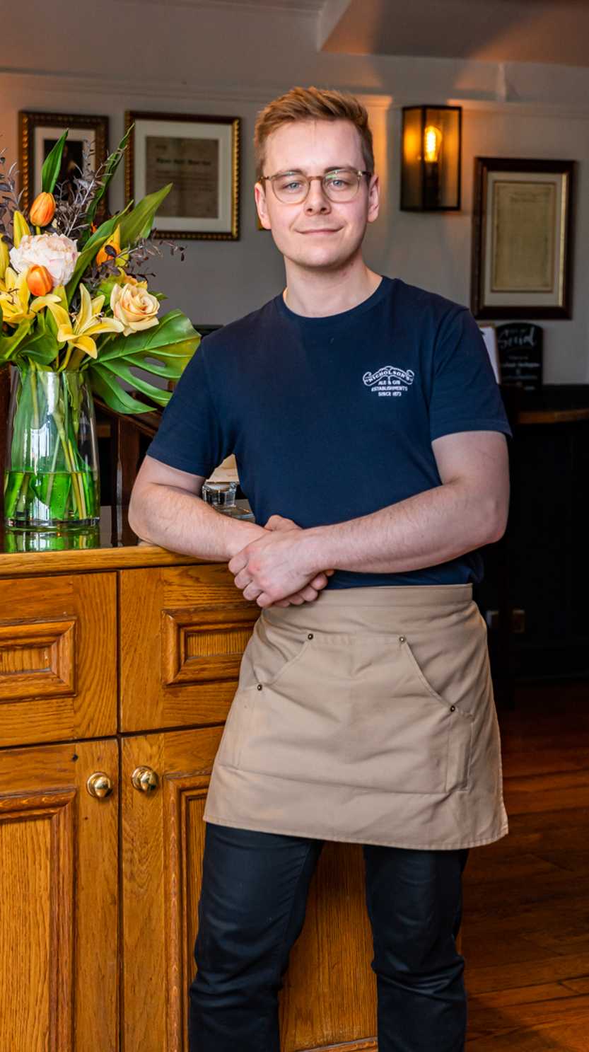 A team member in a blue t-shirt and brown apron leans against a cabinet, with fresh flowers in a vase on top.