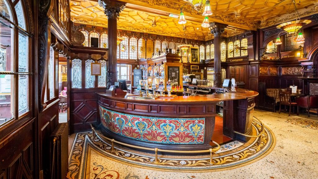 A circular bar stands in the corner of a grand pub, with delicate lamp shades hanging from a detailed ceiling.