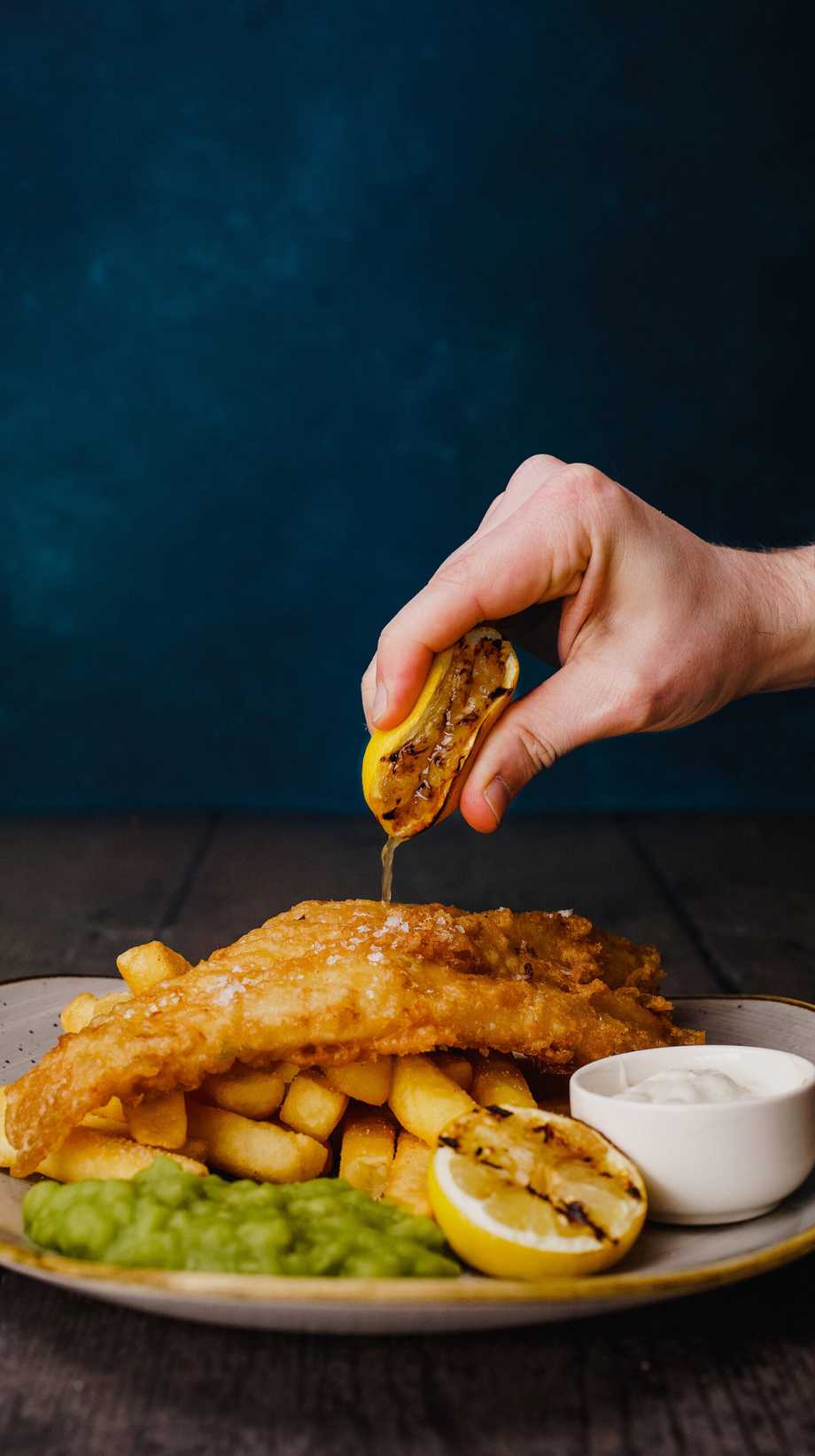 A hand squeezes fresh lemon over a plate of fish and chips, including mushy peas and tartar sauce.