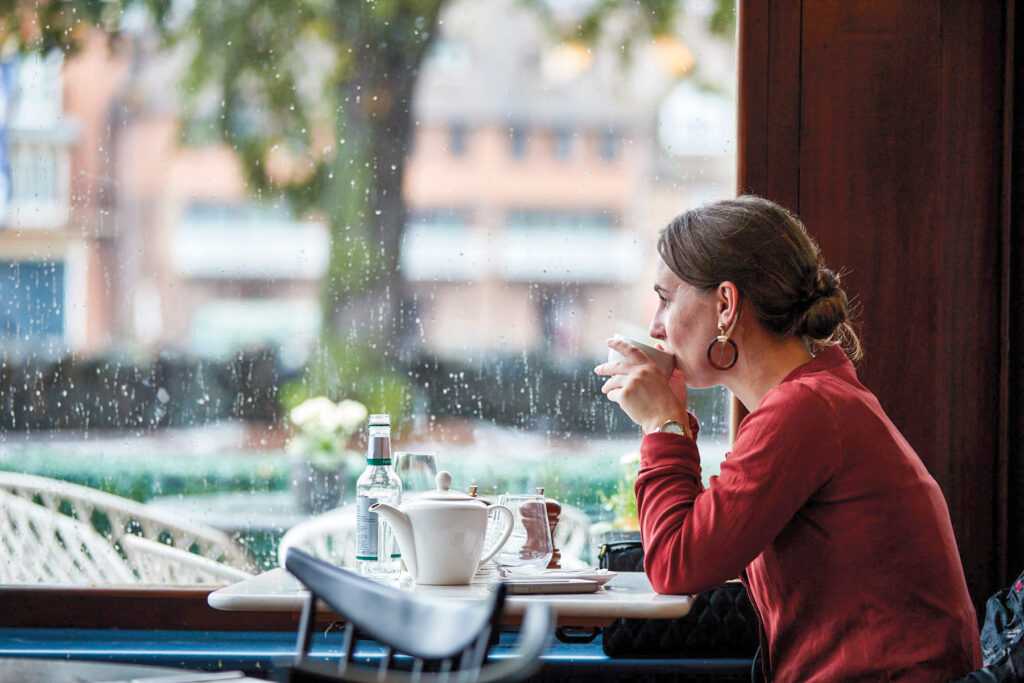 A guest in a red jumper stares out the window while drinking tea. Outside, rain is hitting the window.