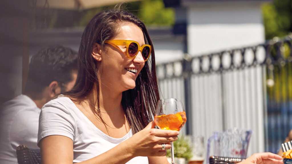 A smiling woman in sunglasses, holding an orange drink in her hand.