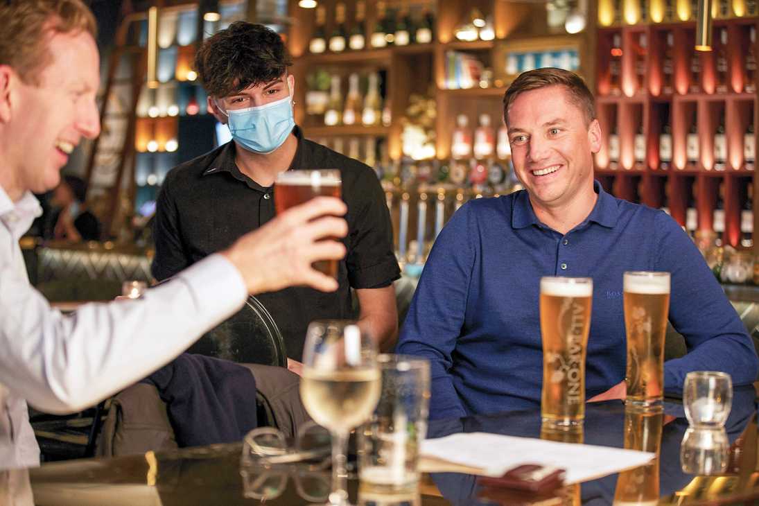Two men are catching up over several pints while a masked team member watches.