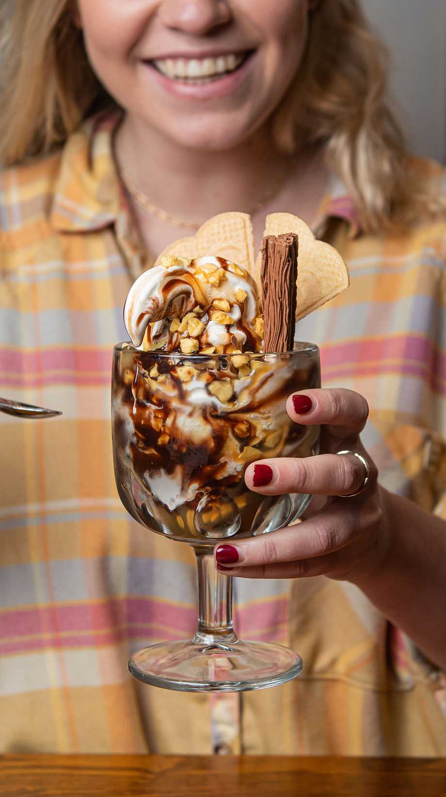 A guest holds a chocolatey sundae with wafers and a flake up to the camera while smiling.