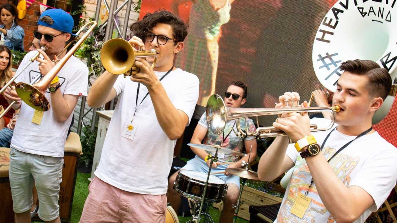 A band is performing. In the foreground, three men play trumpets or trombones. Behind them, a man plays the drums.