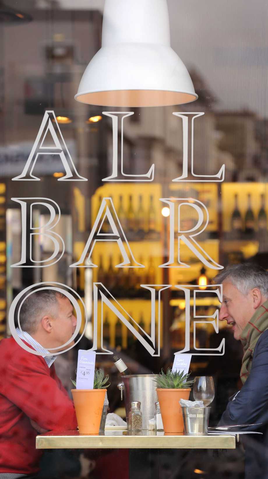 Two men chat at All Bar One, sitting behind the All Bar One logo on the window.