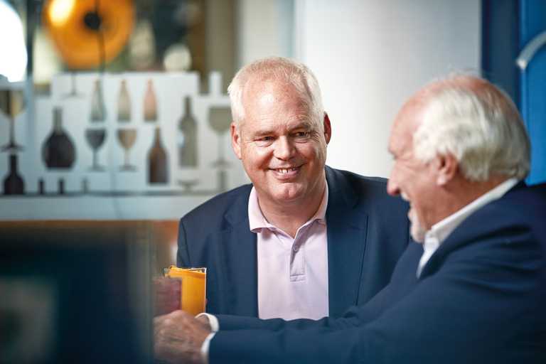 Two men sit in a booth smiling. One is wearing a blue shirt with a pink polo shirt, and the other is wearing a blue suit with a white shirt.