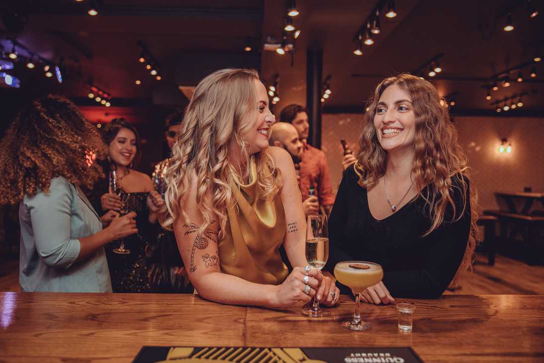 Two women stand at the bar with their drinks, talking. Behind them, the party is in full swing.
