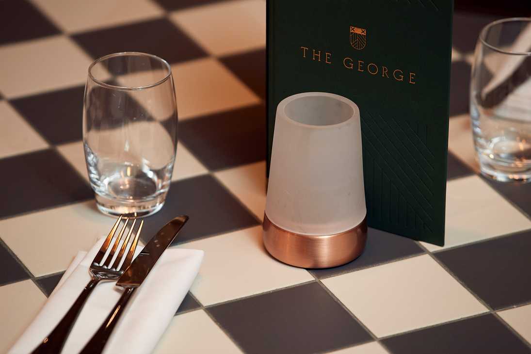 A checkered table is carefully laid at The George of Harpenden pub, including a napkin, cutlery, glass, candle, and menu.