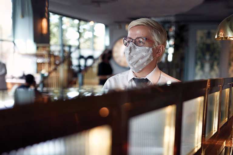 A team member wears a fabric face mask decorated with stars. Running through the centre of the image is a gently lit wall, diving the bar.