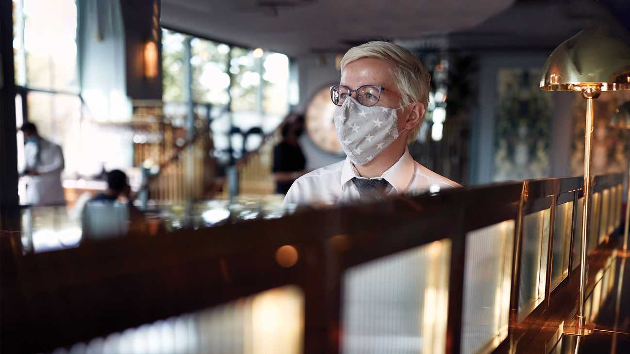 A team member wears a fabric face mask decorated with stars. Running through the centre of the image is a gently lit wall, diving the bar.