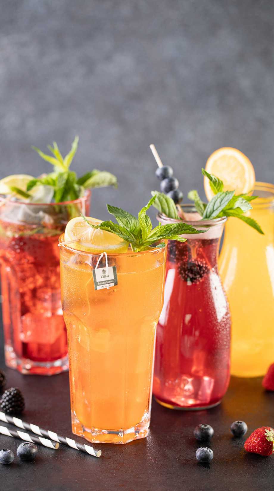 A selection of chilled drinks with fruity garnishes are placed on a table. Around them, striped straws and fresh fruit.