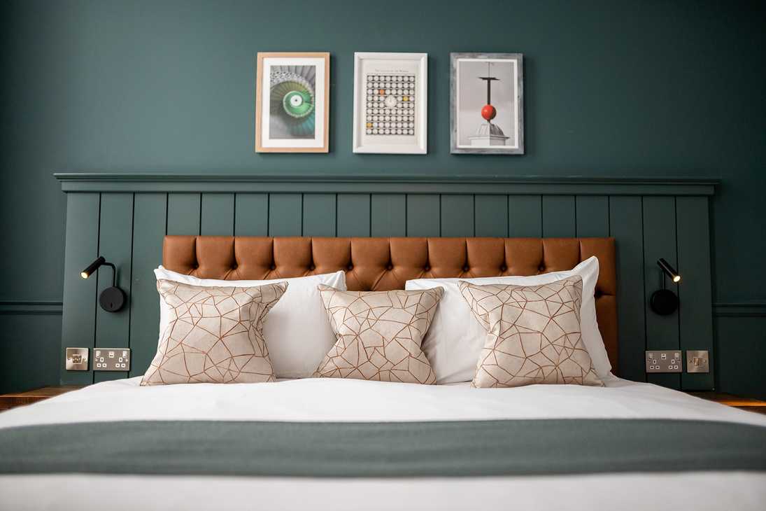 A bed with a white sheet, fluffy cushions, and warm, brown headboard is set against a teal feature wall.