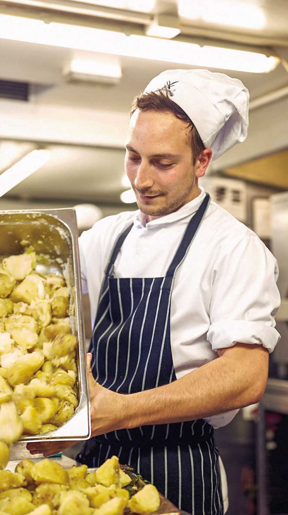 A chef with an apron and white hat pours a tub of roast potatoes into another tub.