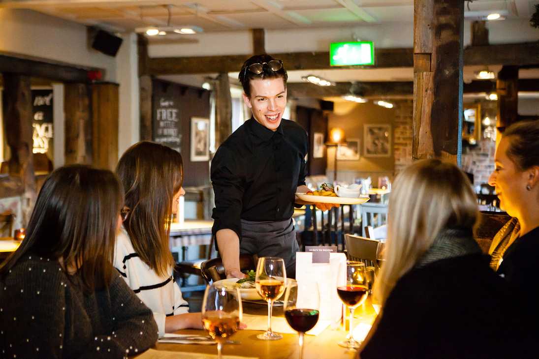 A smiling member serves meals to a table of waiting guests. In front of them are four glasses of wine.