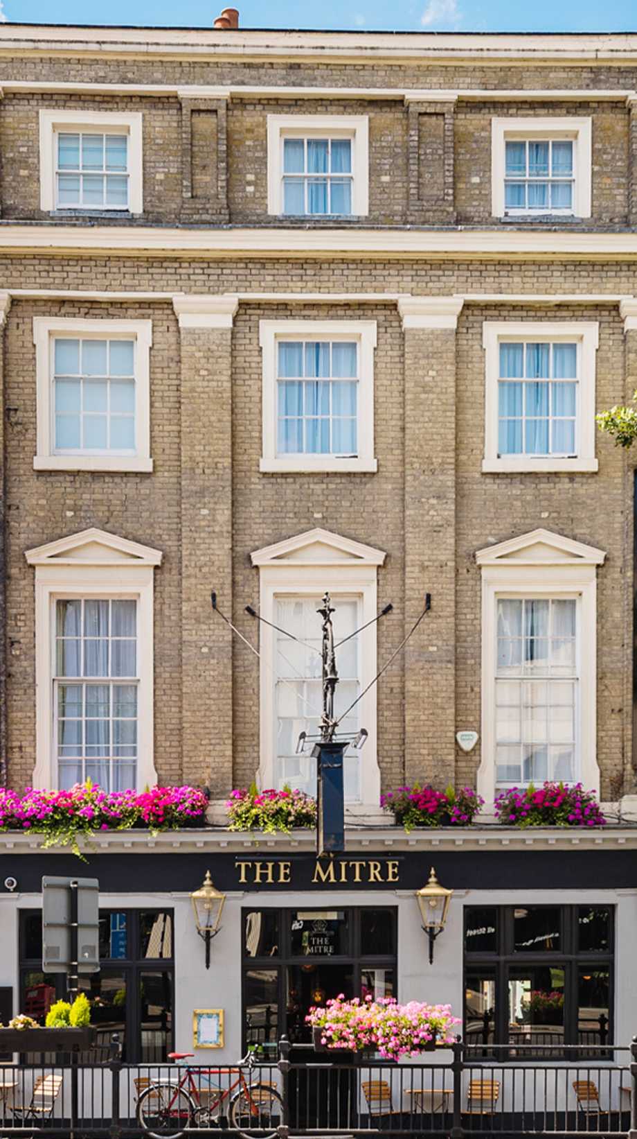 A portrait shot of The Mitre hotel, an Innkeeper's in London. It has high windows and pink window-box flowers with gold lighting adorning the entrance.
