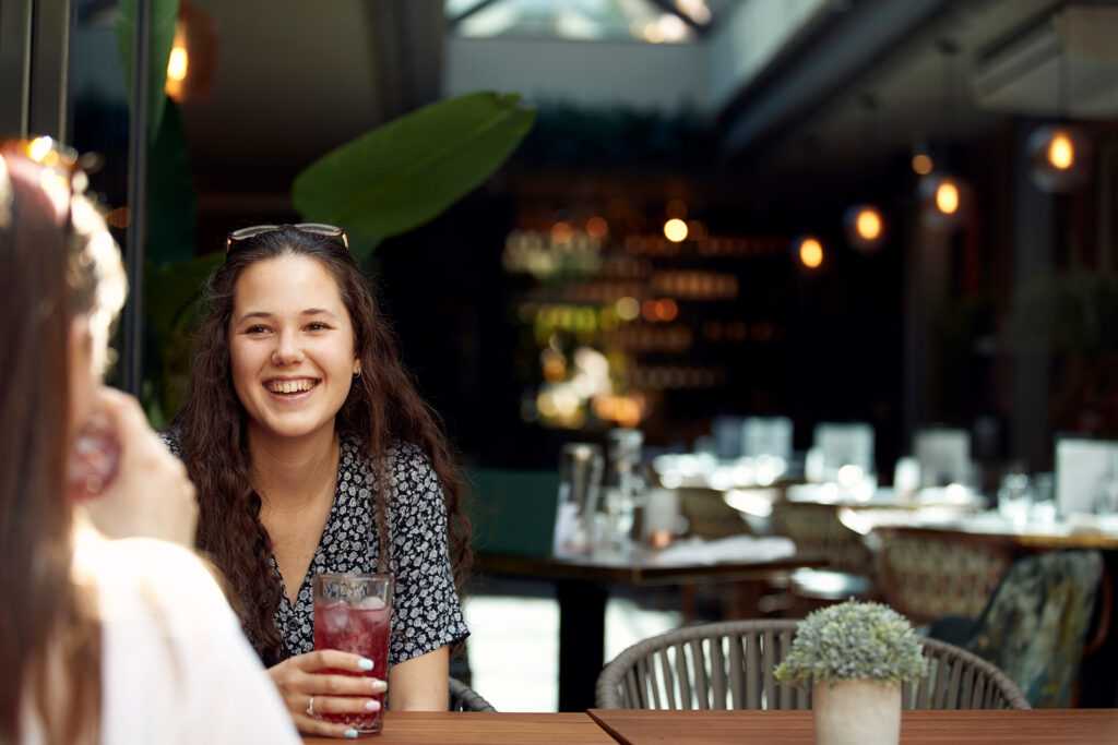 A girl with glasses resting on her head smiles at her friend, sitting in a tastefully decorated bar with plants and exposed lightbulbs.