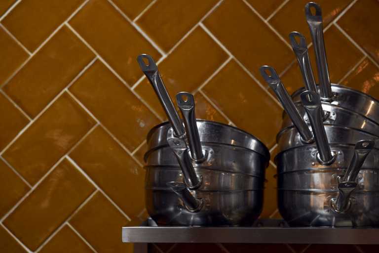 Pots and pans are piled high on a metal shelf, sitting in front of a burnt orange tile.