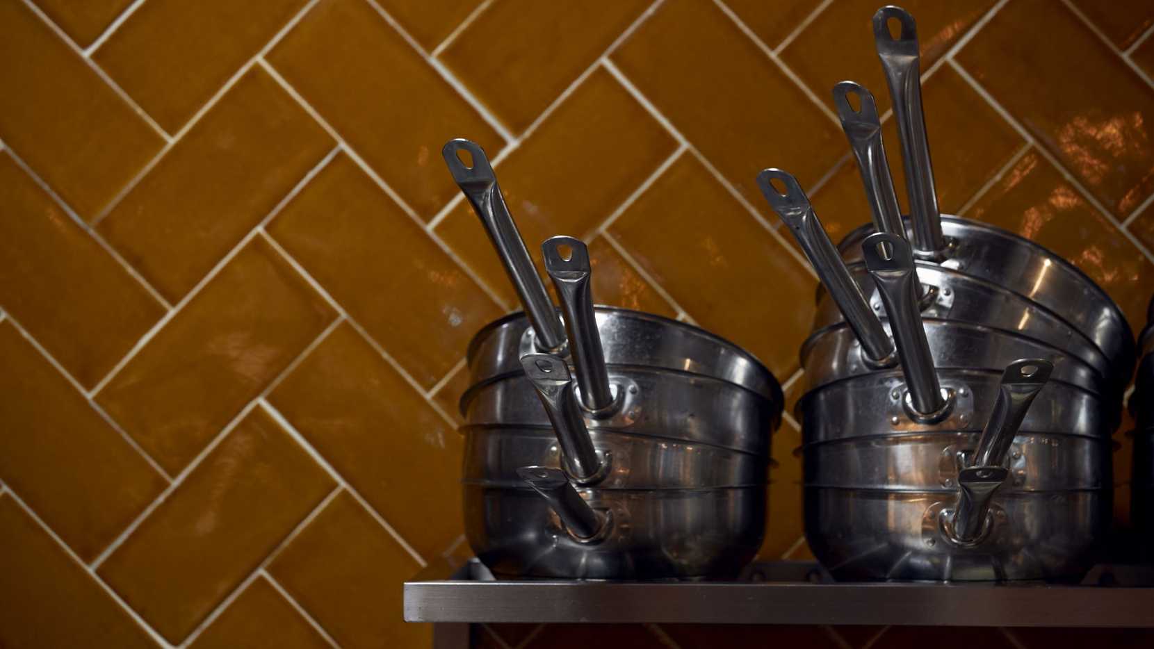 Pots and pans are piled high on a metal shelf, sitting in front of a burnt orange tile.