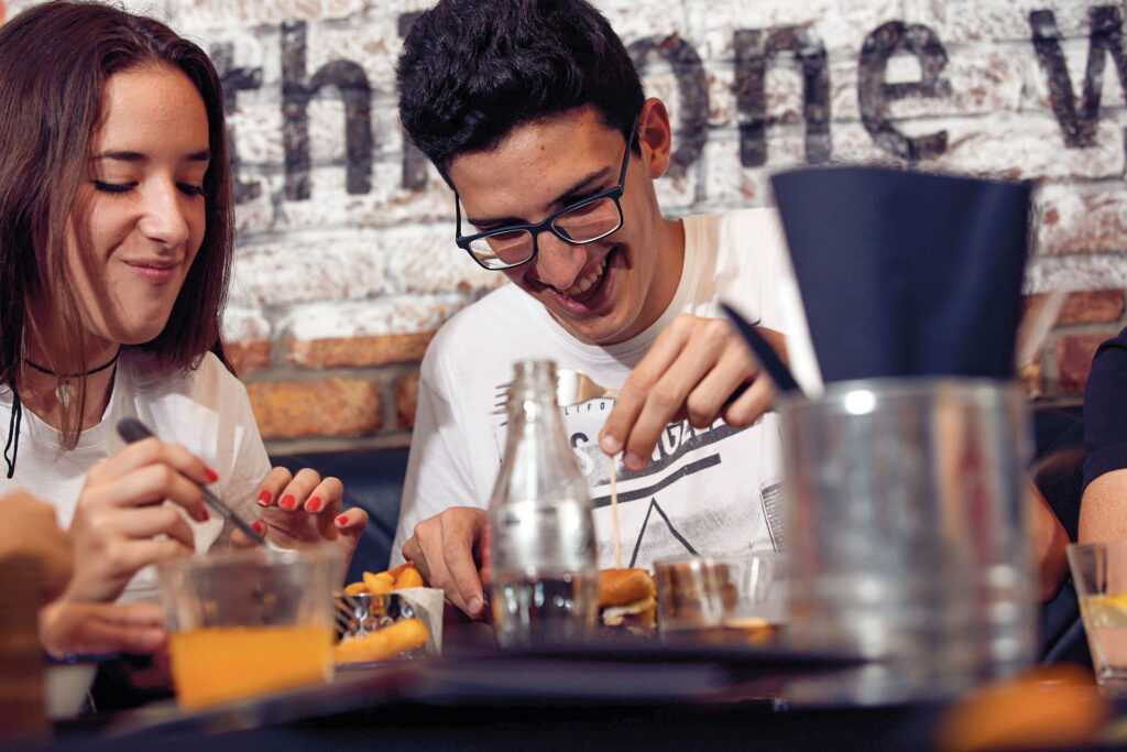 A couple are smiling while eating burgers and chips. The man is removing the toothpick from his bun.