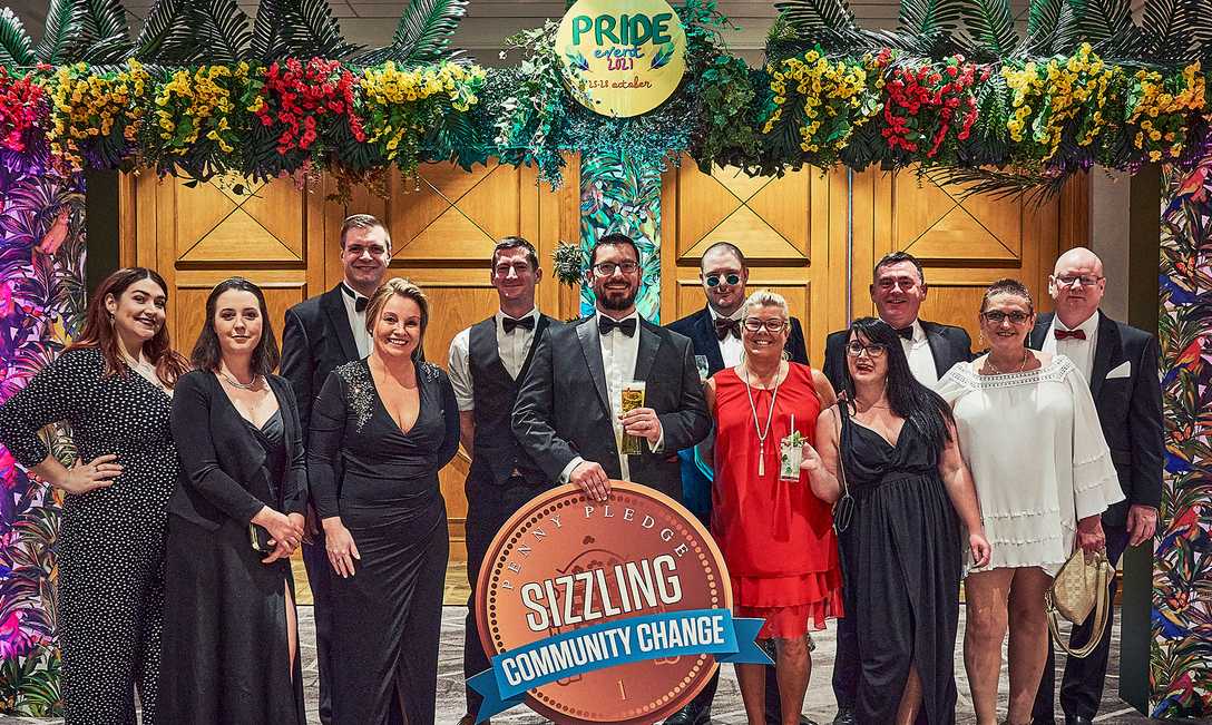 The Sizzling team standing in an arch of flowers for a Pride 2021 event, holding a massive coin-shaped sign that says, "Penny Pledge: Sizzling Community Change."