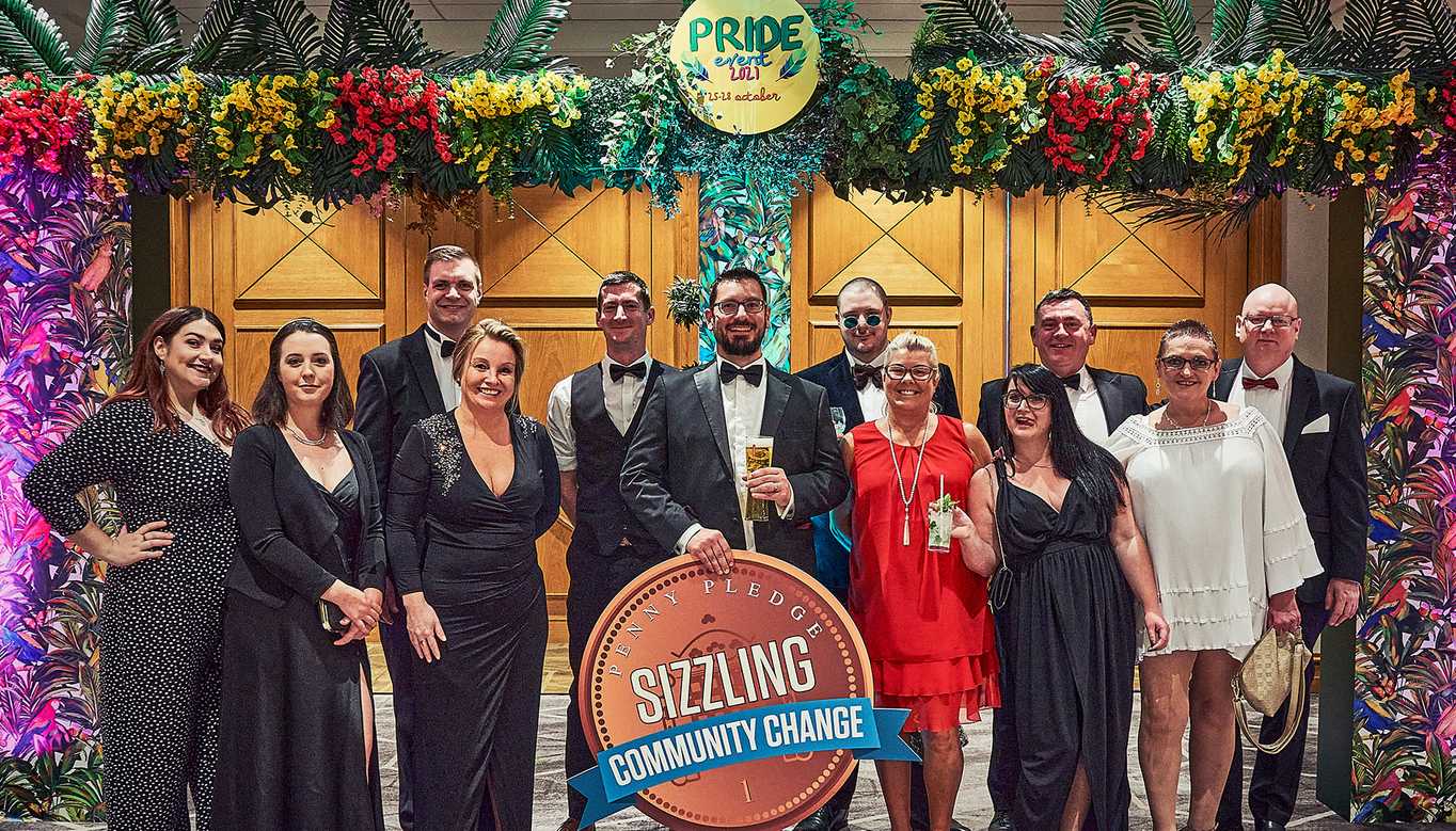 The Sizzling team standing in an arch of flowers for a Pride 2021 event, holding a massive coin-shaped sign that says, "Penny Pledge: Sizzling Community Change."