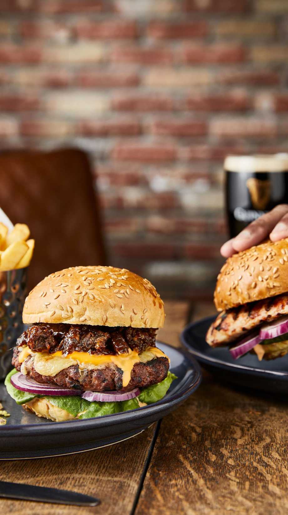 A hand is picking up a large burger. Next to it, there's another burger, chips, and a pint of Guinness.