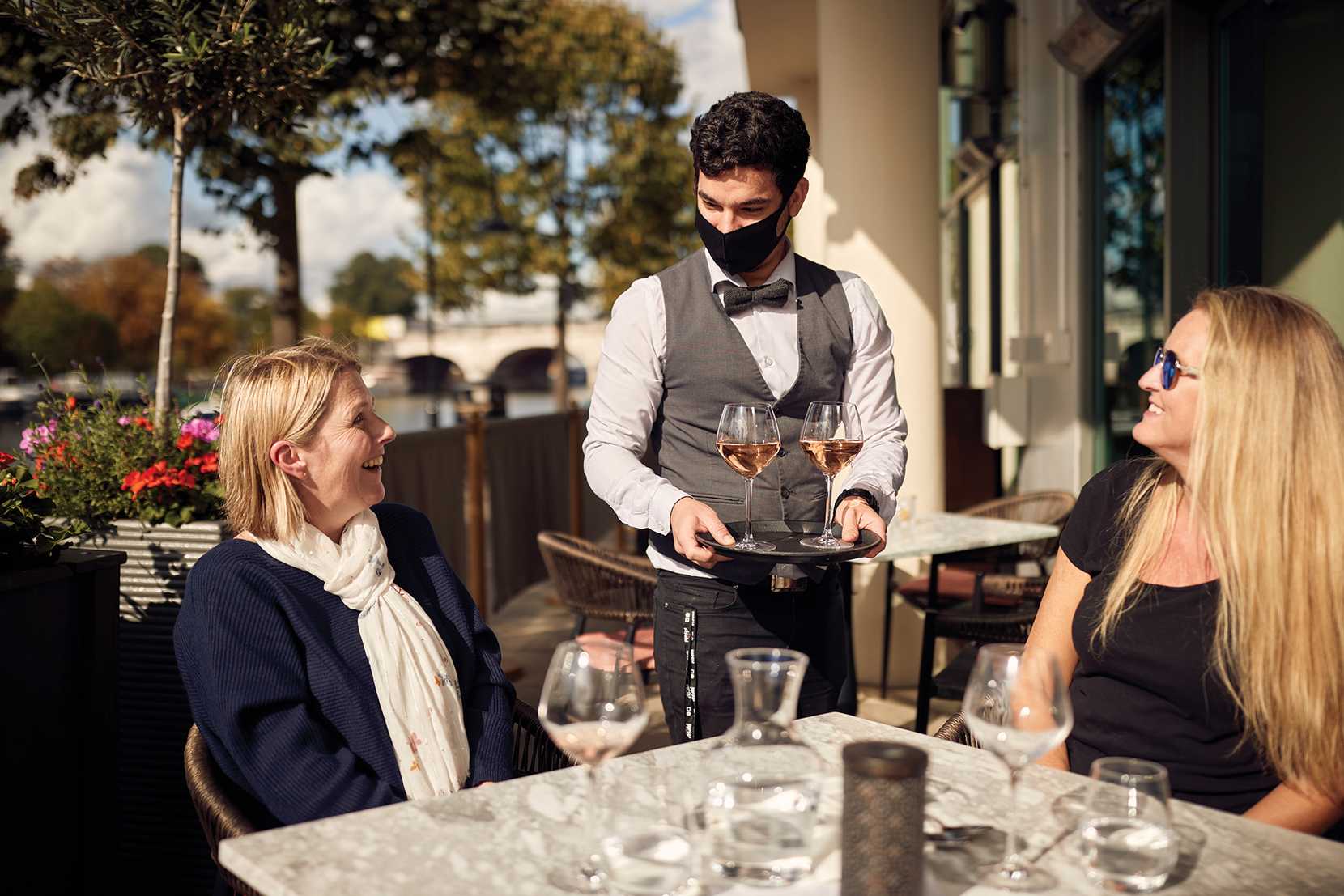 A team member wearing a face mask, waistcoat, and bowtie serves two glasses of wine to waiting guests, sitting in the sun.