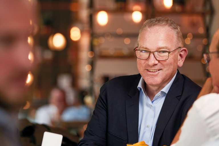 A man in a dark blue suit and light blue shirt smiles while holding his drink.