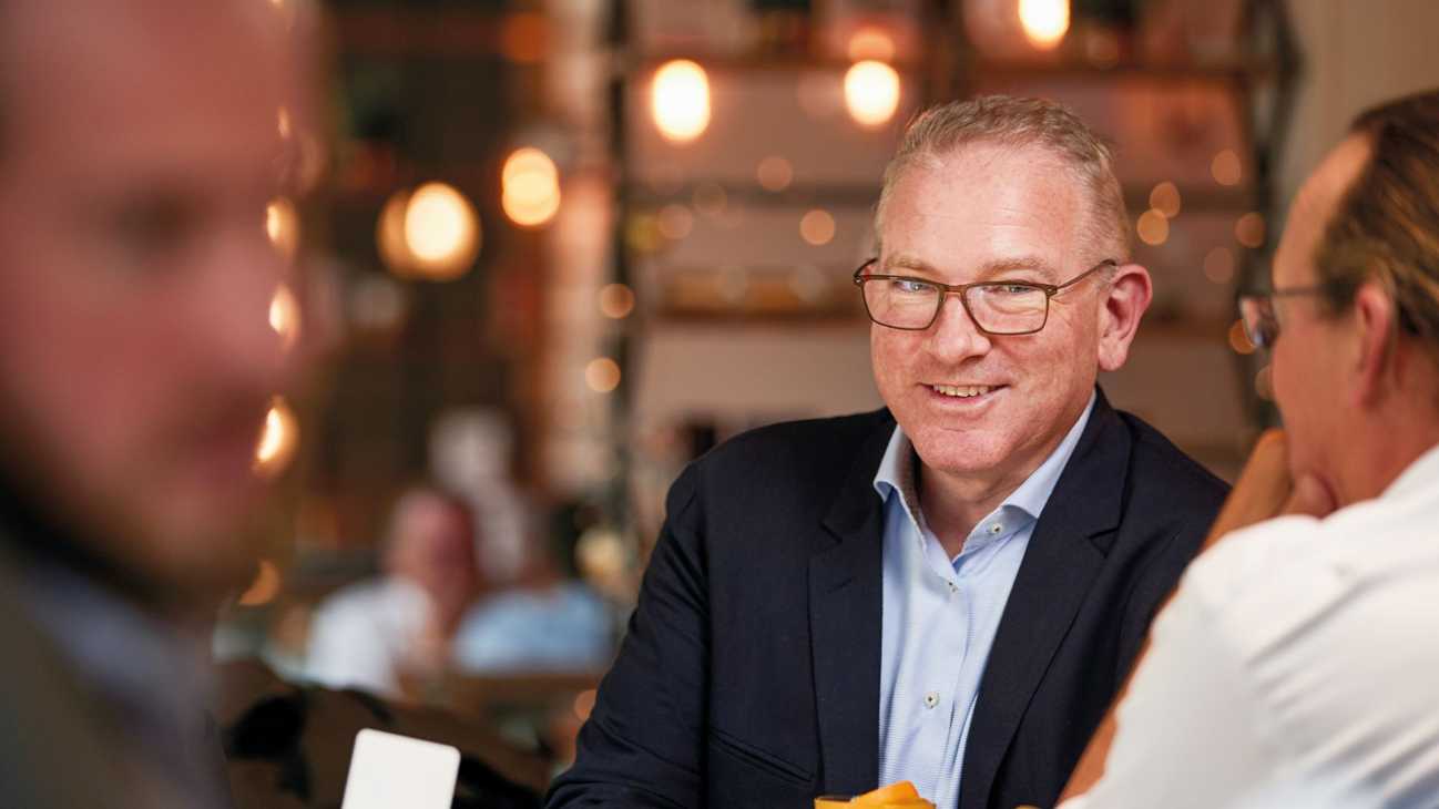 A man in a dark blue suit and light blue shirt smiles while holding his drink.